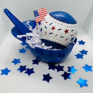 May include: A white baseball with blue and red star and dash patterns sits inside a blue plastic mini helmet bowl filled with white paper shred. A small American flag on a toothpick is inserted into the bowl, surrounded by blue star confetti.