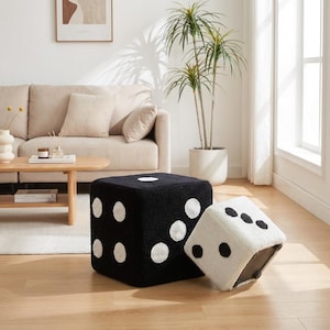 May include: Two dice-shaped ottomans, one black with white dots and the other white with black dots, sit on a hardwood floor. The black ottoman is upright, while the white one is tilted. The background includes a beige sofa, a coffee table, and a potted plant.