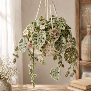 May include: A crocheted hanging plant featuring multiple shades of green monstera-style leaves and small pink and white flowers. The plant is contained within a cream-colored crocheted basket suspended by four thick, braided off-white cords.