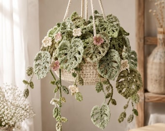 Puede incluir: Una planta colgante de ganchillo con hojas tipo monstera en varios tonos de verde y pequeñas flores rosas y blancas. La planta está contenida en una cesta de ganchillo color crema suspendida por cuatro cordones gruesos trenzados.