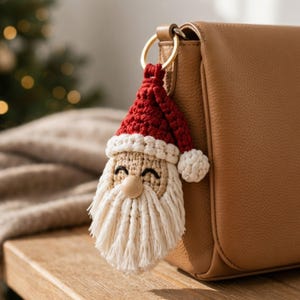 Könnte beinhalten: Ein gehäkelter Weihnachtsmann-Schlüsselanhänger mit einer roten Mütze und weißem Bommel, einem beigen Gesicht mit schwarzen gestickten Augen, einer glatten Holzkugelnase und einem langen, fransigen weißen Bart an einem goldfarbenen Metallring.