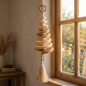 May include: A hanging spiral ornament crafted from cream-colored cord, featuring a conical shape that tapers toward the bottom. The structure is suspended from a wooden ring and finished with a decorative tassel and small wooden beads at the base.