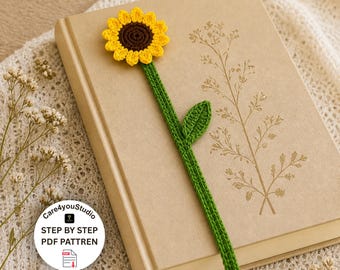 Puede incluir: Un marcapáginas de girasol tejido a ganchillo con una flor amarilla, centro marrón y tallo y hoja verdes. El marcapáginas descansa sobre un libro cerrado con ilustraciones florales. El texto "STEP BY STEP PDF PATTREN" es visible.