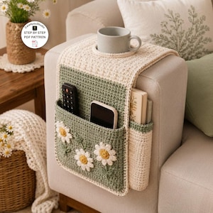 Pode incluir: Um organizador de braço de sofá em crochê verde-sálvia e creme, com três aplicações de flores margaridas brancas, um porta-copos integrado e vários bolsos contendo um controle remoto preto, um smartphone e um livro.