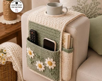 Puede incluir: Un organizador de ganchillo para reposabrazos de sofá en color verde salvia y crema, con tres aplicaciones de flores de margarita blancas, un portavasos integrado y varios bolsillos con un mando a distancia negro, un smartphone y un libro.