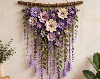 Puede incluir: Un tapiz de pared con flores tejidas a ganchillo en tonos morado, lavanda y crema, acentuado con hojas y enredaderas verdes tejidas. El arreglo floral cuelga de una rama de madera natural y presenta borlas decorativas en color morado claro.