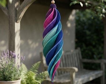 Puede incluir: Un adorno colgante en espiral hecho de hilo de ganchillo en un degradado de colores vibrantes, que transita desde el magenta intenso y el morado hasta el turquesa y el azul. La pieza tiene una cuenta de madera arriba y un cristal morado abajo.