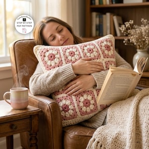 May include: A square crochet pillow featuring a repeating pattern of dusty rose and cream-colored floral granny squares. The pillow is constructed from joined squares with a textured yarn finish, showcasing a consistent geometric design throughout.