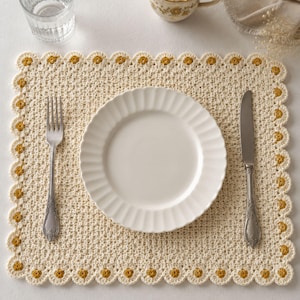 May include: A rectangular cream-colored crocheted placemat featuring a scalloped border with small mustard yellow flower accents. A white fluted ceramic plate sits in the center, flanked by a silver fork on the left and a silver knife on the right.