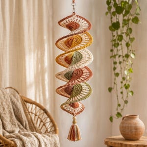 May include: A hanging spiral decoration crafted from crocheted yarn in shades of rust, mustard yellow, sage green, and cream. The structure features a repeating fan-like pattern and terminates in a wooden bead and a multi-colored yarn tassel.