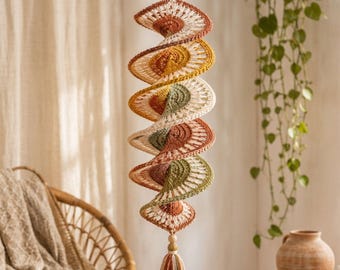 Puede incluir: Una decoración en espiral colgante hecha de hilo de ganchillo en tonos óxido, amarillo mostaza, verde salvia y crema. La estructura presenta un patrón repetitivo en forma de abanico y termina en una cuenta de madera y una borla de hilo multicolor.