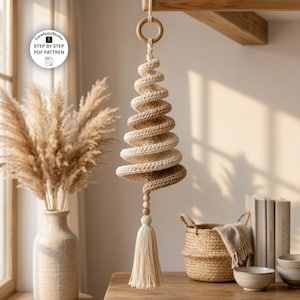 Op de afbeelding: Een hangend spiraalvormig kerstboomornament gemaakt van crème en beige garen. De structuur heeft een houten ring aan de bovenkant en twee kleine houten kralen boven een lange, dikke, crèmekleurige kwast. De tekst luidt Care4youStudio STEP BY STEP PDF PATTREN.