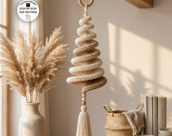 Puede incluir: Un adorno colgante de árbol de Navidad en espiral hecho de hilo color crema y beige. La estructura cuenta con un anillo de madera en la parte superior y dos pequeñas cuentas de madera sobre una borla larga y gruesa de color crema. El texto dice Care4youStudio STEP BY STEP PDF PATTREN.