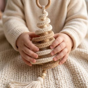 May include: A spiral-shaped crochet ornament featuring alternating bands of cream and tan yarn. The ornament hangs from a smooth wooden ring at the top and terminates in a small wooden bead and a cream-colored yarn tassel at the bottom.