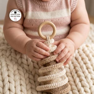 May include: A spiral-shaped crochet toy featuring a natural wooden ring at the top, crafted in alternating shades of cream and beige yarn. The circular text on the image reads Care4youStudio STEP BY STEP PDF PATTREN with a document icon.