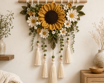 Puede incluir: Un tapiz de pared que presenta un gran girasol central de ganchillo rodeado de margaritas blancas más pequeñas, hojas verdes y bayas. El arreglo cuelga de un soporte de madera con borlas de hilo color crema y cuentas de madera colgantes.
