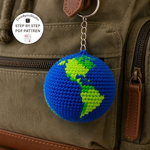 May include: A blue and green crocheted globe keychain. The continents are in green, and the oceans are blue. The keychain is attached to a silver chain and a metal ring. The background is a green backpack.