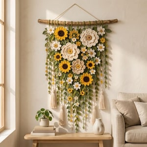 May include: A wall hanging featuring a collection of crocheted sunflowers and white daisies attached to a horizontal wooden branch. The arrangement includes green crocheted leaves and vines, with several cream-colored tassels hanging from the bottom.