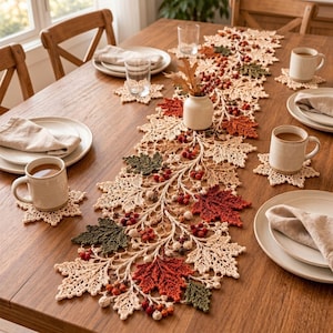 Pode incluir: Um longo caminho de mesa em crochê com um padrão intrincado de folhas de bordo na cor creme, intercalado com folhas de crochê em vermelho vibrante e verde escuro e pequenos cachos de bagas, acompanhado por porta-copos a condizer em forma de folha.