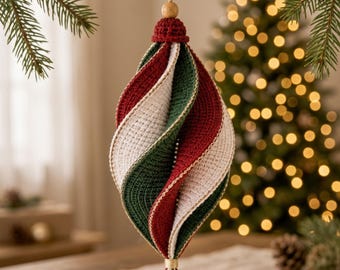 Puede incluir: Un adorno navideño en forma de espiral hecho de hilo de ganchillo con segmentos alternos en rojo oscuro, verde bosque y blanco crema. El adorno cuenta con una pequeña cuenta de madera en la parte superior, un cordel y una borla decorativa abajo.