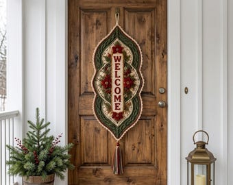 Puede incluir: Un tapiz vertical alargado de ganchillo con un panel central color crema que muestra la palabra WELCOME en letras rojas, rodeado de bordes de punto trenzado verde oscuro, detalles de flores de pascua rojas y una borla multicolor.