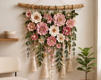 Puede incluir: Un tapiz de pared con flores de ganchillo en tonos rosa, blanco y crema, intercaladas con hojas y enredaderas de ganchillo verdes. El arreglo cuelga de una rama de madera natural con borlas de hilo de color crema colgando debajo.