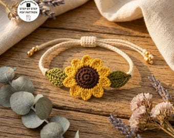 Puede incluir: Una pulsera de ganchillo con un girasol amarillo central de centro marrón oscuro y dos hojas verdes a cada lado. La pulsera tiene un cordón de color crema con un cierre de nudo deslizante y pequeñas cuentas doradas en los extremos.