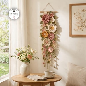 May include: A vertical wall hanging featuring a collection of crocheted flowers and leaves in shades of dusty rose, cream, and mustard yellow, suspended from a wooden dowel with a string, with text reading Care4youStudio STEP BY STEP PDF PATTREN.