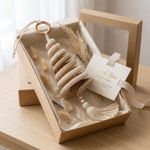 May include: A spiral-shaped crochet ornament in shades of beige and brown, featuring a wooden ring at the top and a tassel at the bottom. It is presented in a cardboard gift box with dried grasses and a card that reads Thank You for your order.