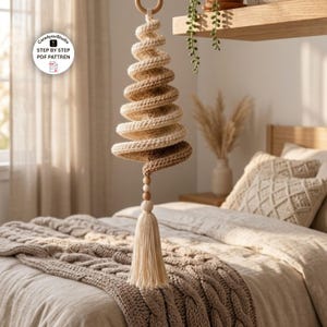 May include: A hanging spiral crochet ornament in cream and tan yarn, suspended from a wooden ring. The structure features a tapered, coiled design ending in two wooden beads and a long, cream-colored tassel. Text reads: Care4youStudio STEP BY STEP PDF PATTREN.