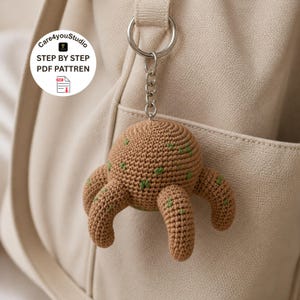 Op de afbeelding: Een gehaakte bruine sleutelhanger in de vorm van een wezen met vier poten en groene accenten. De sleutelhanger is bevestigd aan een zilveren ring en ketting. De tekst "Care4youStudio STEP BY STEP PDF PATTREN" staat in een witte cirkel.