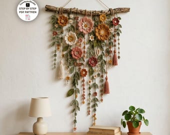May include: A wall hanging featuring crocheted flowers and leaves in shades of rust, pink, and green, suspended from a natural wooden branch. The piece includes hanging vines, small blossoms, and decorative tassels, with a circular logo in the corner.