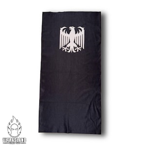 Könnte beinhalten: Schwarzes rechteckiges Textil mit weißem deutschem Adler-Emblem. Das Textil hat ein strukturiertes Aussehen und ist wahrscheinlich aus Stoff. Das Logo von ULTRASLAND GERMAN SHOP ist unten links zu sehen.
