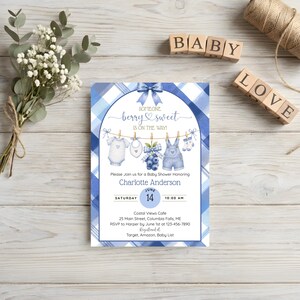 May include: A rectangular baby shower invitation card featuring a blue plaid border, a central arch, and a clothesline with a onesie, bib, blueberries, overalls, and socks. The text reads Someone berry sweet is on the way, Charlotte Anderson, June 14.