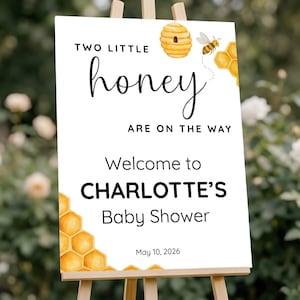 May include: A white rectangular sign featuring the text "TWO LITTLE honey ARE ON THE WAY" and "Welcome to CHARLOTTE'S Baby Shower" with the date "10 May 2026". The design includes watercolour illustrations of a beehive, a bee, and yellow honeycombs.