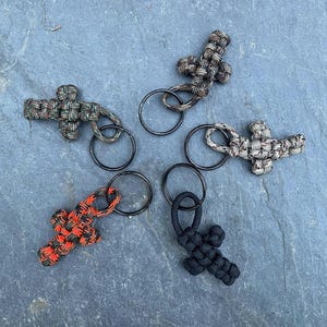 May include: Five keychains featuring cross-shaped charms woven from paracord. The collection includes one solid black cross, one bright orange and black patterned cross, and three crosses in various camouflage patterns of green, brown, and tan.