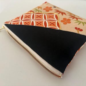 May include: A square, patterned pouch with a gold zipper. The pouch features a black panel and a beige panel with orange floral and geometric designs. The zipper pull is gold.