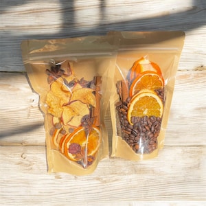 May include: Two clear plastic bags filled with dried fruit and spices. One bag contains dried apple slices, orange slices, star anise, and cinnamon sticks. The other bag has orange slices, coffee beans, and cinnamon sticks. The bags are on a light wood surface.