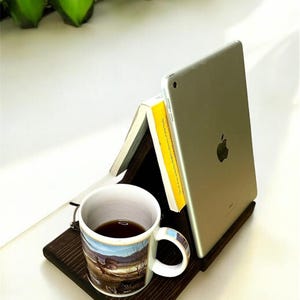 May include: A dark brown wooden tray holds a coffee mug, a book, and a silver tablet. The mug has a landscape design. Green plant-like decorations are in the background. The tablet has an Apple logo.