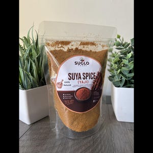 Könnte beinhalten: Ein transparenter Plastikbeutel gefüllt mit Suya Spice (Yaji) von Suglo Foods. Das Etikett zeigt Text und eine Abbildung des Gewürzes und des gegrillten Fleisches. Das Gewürz wird als 100% natürlich beschrieben, mit einer authentischen Erdnuss- und Pfeffermischung.