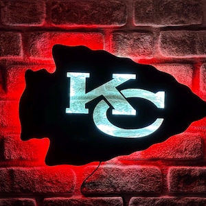 May include: A black arrowhead-shaped sign with the letters "KC" in white, illuminated against a red brick wall. The sign is lit with a soft glow, and a power cord is visible.