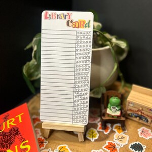May include: A white library card with the words "Library Card" in colorful letters. The card has lines for writing and star icons. It is displayed on a small wooden easel, surrounded by leaf stickers and a miniature scene.
