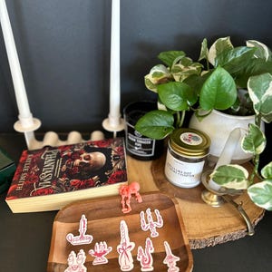 Può includere: Una composizione natura morta con un vassoio di legno con adesivi di candele rosa, un libro intitolato "Phantasma" e due candele affusolate bianche in supporti. Sono presenti anche una piccola pianta in vaso e una candela in un barattolo, creando una scena decorativa e accogliente.
