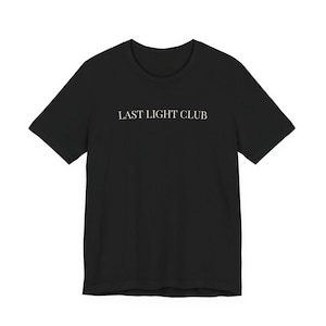 May include: Black t-shirt with the text "LAST LIGHT CLUB" in a simple, sans-serif font across the chest. The shirt has short sleeves and a crew neck. The fabric appears to be a soft, opaque material.