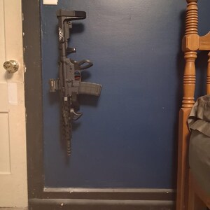 May include: A black and gray rifle mounted on a dark blue wall. The rifle has a magazine, a handle, and a stock. The wall has a light switch and is next to a white door and a wooden bed frame.