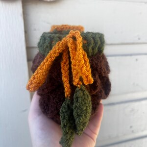 May include: A small, round, crocheted pouch in dark brown yarn with a dark green top border. The pouch features a drawstring closure made of orange yarn, ending in two small, crocheted green leaf-shaped accents hanging from the front.