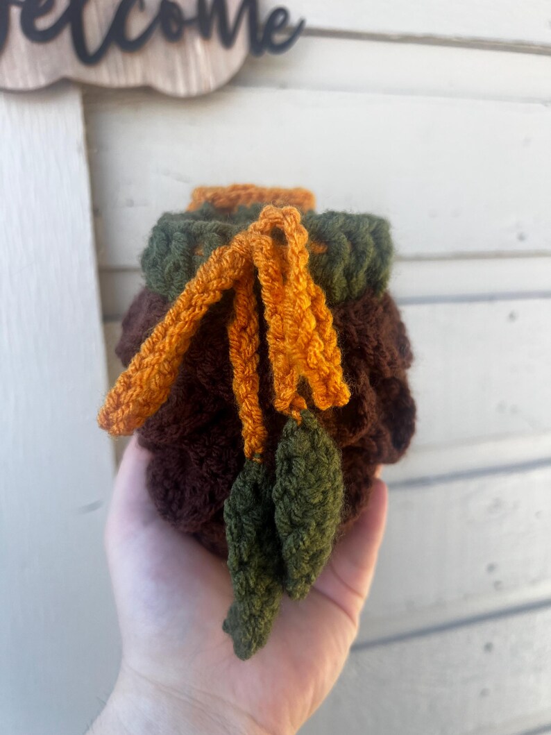 May include: A small, round, crocheted pouch in dark brown yarn with a dark green top border. The pouch features a drawstring closure made of orange yarn, ending in two small, crocheted green leaf-shaped accents hanging from the front.