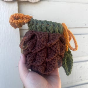 May include: A crochet pouch shaped like a pinecone with a dark brown textured body, a dark green ribbed top, and an orange drawstring closure. A small brown leather tag is attached to the side, and a single dark green crocheted leaf is attached.