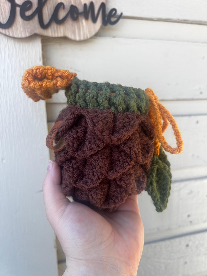 May include: A crochet pouch shaped like a pinecone with a dark brown textured body, a dark green ribbed top, and an orange drawstring closure. A small brown leather tag is attached to the side, and a single dark green crocheted leaf is attached.