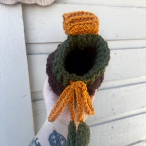 May include: A crochet drawstring pouch featuring a dark brown base, a forest green upper section, and a bright orange top flap. The pouch includes an orange drawstring cord finished with two small, dark green crocheted leaf-shaped accents.